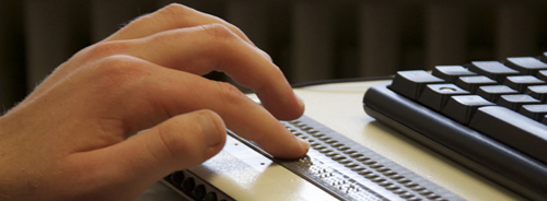 Vor der Computertastatur liegt eine Braille-Zeile zum Auslesen der Brailleschrift; mit einer Händen liest eine Person gerade einen Text. (Foto: Birgit Guggi)