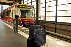 Das Foto zeigt einen blinden Mann mit Langstock auf einem Bahnsteig, der auf ein dort abgestelltes Gepäckstück zugeht.